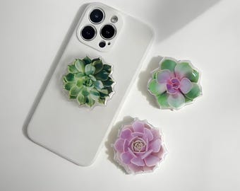 Peut inclure: Trois supports de téléphone en forme de succulentes, dans des tons de vert et de violet. L'un est fixé à un téléphone blanc. Les deux autres sont placés séparément. Les supports ont une bordure transparente et texturée.