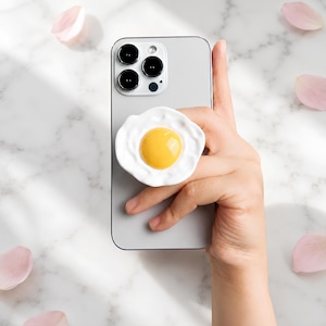 Op de afbeelding: Een grijze smartphone met een ronde telefoonhouder aan de achterzijde, ontworpen als een gebakken ei met een glanzende, verhoogde gele dooier in het midden en een onregelmatige, getextureerde witte rand voor een stevige grip op het toestel.