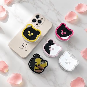 Könnte beinhalten: Eine beige Smartphone-Hülle mit dem Text "good luck" und einem Smiley-Gesicht, begleitet von vier bärenförmigen Handyhaltern. Die Halter haben reflektierende Spiegelflächen mit gelben, rosa, schwarzen und weißen Umrandungen und Bärengesichtern.