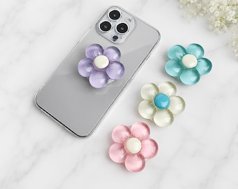 Peut inclure: Un smartphone transparent avec un support en forme de fleur violette fixé au dos, accompagné de trois autres supports en forme de fleur bleu sarcelle, blanc translucide avec un centre bleu, et rose, chacun avec un centre circulaire blanc ou bleu.