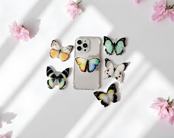 May include: A collection of butterfly-themed phone grips and a clear phone case, arranged on a white surface with pink flowers. The butterfly grips feature various colors and patterns, including yellow, blue, and green. The scene is lit with natural light.