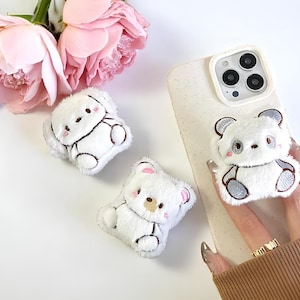 May include: White plush bear phone grip and two other plush bear accessories. The bear accessories have black embroidered outlines and pink accents. A white phone case with a bear grip is also in the image, along with pink roses.