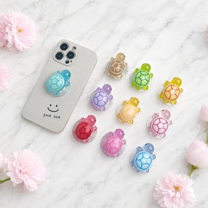 May include: A selection of colourful turtle-shaped phone grips in shades of blue, green, pink, and yellow. One is attached to a light grey phone case with a smiley face and the words "good luck". Pink flowers are scattered around the grips.