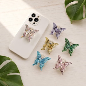 Könnte beinhalten: Ein weißes Smartphone mit einem bunten, mit Strasssteinen besetzten Schmetterlings-Handygriff auf der Rückseite, begleitet von fünf weiteren Schmetterlings-Handygriffen in Lila, Gold, Grün, Blau und Pink, die jeweils mit funkelnden Strasssteinen verziert sind.