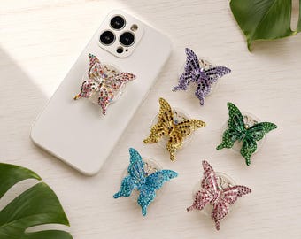 May include: A white smartphone with a multi-colored rhinestone butterfly phone grip attached to the back, accompanied by five additional butterfly phone grips in purple, gold, green, blue, and pink, each encrusted with sparkling rhinestones.