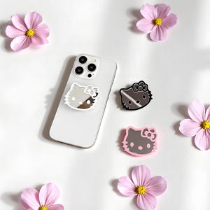 Könnte beinhalten: Ein weißes Smartphone mit einem silbernen Hello Kitty-Handyhalter, zusammen mit zwei zusätzlichen Hello Kitty-Haltern in Schwarz und Rosa. Rosafarbene Blumen mit gelben Zentren sind um das Telefon herum verstreut und schaffen eine verspielte, feminine Ästhetik.