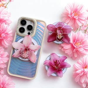 Puede incluir: Una funda de teléfono con un diseño floral y una flor decorativa adjunta. La funda tiene un borde color crema con un anillo de pedrería alrededor de la cámara. También se ven dos agarres para teléfono con forma de flor y flores rosas.