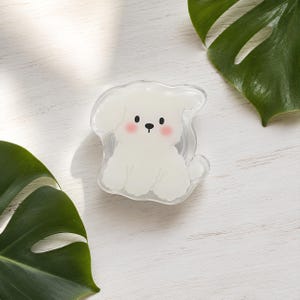May include: A clear, circular phone grip featuring a white, cartoon-style dog illustration. The dog has floppy ears, a small black nose, black eyes, and soft pink circular patches on its cheeks. The design is encased in a transparent acrylic layer.