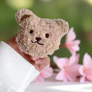 May include: A small, tan teddy bear head with embroidered details. The bear's head is held up against a blurred backdrop of pink flowers and foliage. The bear has dark red eyes and a stitched mouth. The background is out of focus.