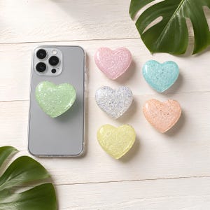 Op de afbeelding: Een smartphone met een lichtgroene hartvormige grip aan de achterkant, vergezeld van vijf extra hartvormige grips in roze, blauw, zilver, perzik en geel. Elk hart heeft een glinsterende, getextureerde afwerking.