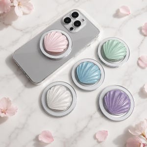 May include: A clear smartphone case with a pink seashell-shaped phone grip attached, accompanied by four additional seashell phone grips in pastel green, blue, white, and purple, each featuring a circular base and a textured, ribbed surface.