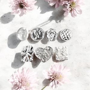 May include: An assortment of silver-toned jewelry pieces, including a coin-shaped pendant, a heart-shaped pendant, and textured rectangular and oval pieces. Pink flowers are scattered around the jewelry on a light-colored surface.