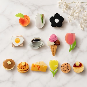 May include: A collection of small decorative pins featuring various food and floral designs, including a peach, a cracked egg, a black flower, toast with a fried egg, a coffee cup, an ice cream cone, a pink tulip, pancakes, a waffle, cheese, a yellow tulip, a cookie, and a scone.