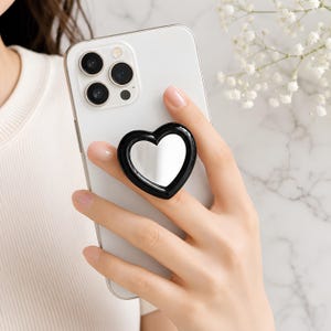 May include: A white smartphone featuring a black heart-shaped phone grip attached to the back. The grip has a glossy black frame surrounding a reflective, mirror-like center surface, providing a functional handle for holding the mobile device.