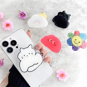 May include: A white smartphone with a white cat-shaped phone grip attached. Other phone grips include a cloud with a yellow star, a black cat, a red blob with eyes, and a colourful flower with a smiley face. Pink and purple flowers are scattered around.