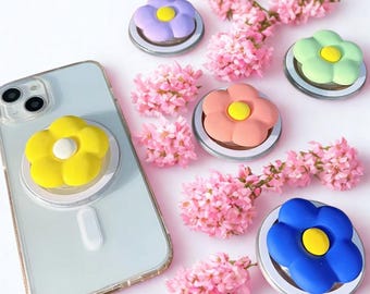 May include: A smartphone with a yellow flower-shaped phone grip attached, surrounded by other flower-shaped grips in purple, green, pink, and blue. Each grip has a yellow center. Pink flowers are scattered around the grips.