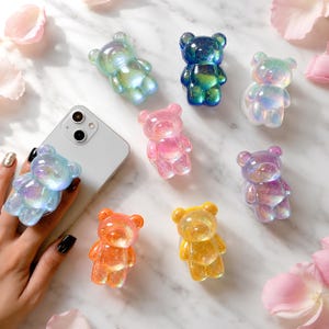 May include: A collection of seven translucent, glittery bear-shaped phone grips in various colors including blue, pink, orange, yellow, and iridescent white. One light blue bear grip is attached to the back of a silver smartphone.