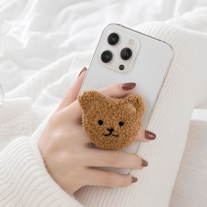 May include: A white smartphone featuring a brown, fuzzy, circular bear-shaped phone grip attached to the back. The bear grip has embroidered black eyes, a nose, and a mouth, with a textured, plush surface that mimics the appearance of soft fur.