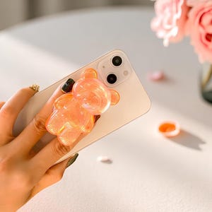 May include: A clear, orange bear-shaped phone grip is attached to a white smartphone. The grip has a glittery appearance. The background includes a vase of pink roses and scattered petals.