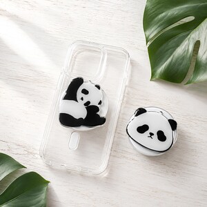 Puede incluir: Una funda de plástico transparente para smartphone con un gráfico de oso panda en blanco y negro en la parte posterior, acompañada de un accesorio de agarre circular para teléfono con forma de cara de panda con orejas, ojos y nariz negros.