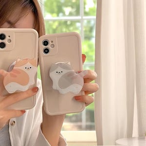 Puede incluir: Dos fundas de teléfono beige con agarres para teléfono con forma de gato. Un agarre para gato es naranja y blanco, el otro es gris y blanco. Las fundas del teléfono tienen una cámara y un flash. El fondo incluye una ventana y una flor rosa.