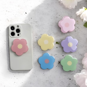 Puede incluir: Un conjunto de agarres para teléfono con forma de flor en colores pastel: rosa, amarillo, morado, azul y verde. Uno está adherido a un teléfono blanco. Cada agarre tiene un centro contrastante. La imagen está sobre un fondo claro con acentos florales.