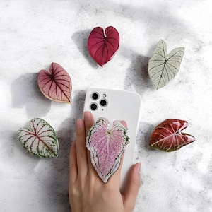 May include: A white smartphone with a leaf-shaped phone grip attached, surrounded by six decorative leaf-shaped phone grips in shades of red, pink, and green. The leaves have intricate vein patterns and are arranged on a white surface.
