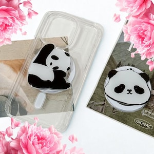 Puede incluir: Funda de teléfono transparente con un diseño de oso panda en blanco y negro. Un agarre para teléfono con forma de oso panda está adherido a la funda. También se ve un agarre para teléfono con forma de oso panda separado. Flores rosas en el fondo.