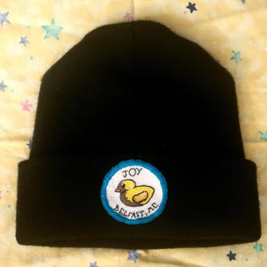 May include: Black beanie hat with a circular patch featuring a yellow duck illustration. The patch has a blue border and the words "JOY BELFAST, ME." The hat is made of a soft, knitted material.