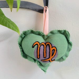 May include: A heart-shaped, green fabric ornament with a ruffled edge. The center features a purple circle with an embroidered orange Virgo symbol. A pink ribbon is attached for hanging.