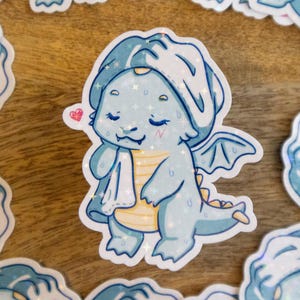 Könnte beinhalten: Ein Aufkleber mit einem niedlichen Zeichentrickdrachen in einem Spa-Setting. Der Drache ist hellblau mit einem weißen Handtuch auf dem Kopf und einem gelben Bauch. Ein kleines rosa Herz befindet sich neben dem Drachen. Andere Aufkleber mit dem gleichen Design umgeben den Hauptaufkleber.