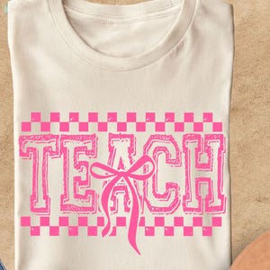 May include: Cream-colored t-shirt with the word "TEACH" in pink, featuring a pink bow and a pink checkered pattern. The shirt is displayed with blue jeans and a brown hat.