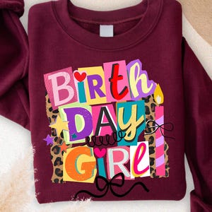 May include: Maroon sweatshirt with the text "BIRTHDAY GIRL" in colorful block letters. The design includes stars, leopard print, and a candle graphic. The sleeves are cuffed.