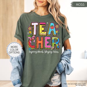 May include: A moss green t-shirt with the word "TEACHER" in colourful, playful lettering. The shirt features a design with a strawberry, pencil, and stars. The text "Inspiring Minds, Shaping Futures" is below the word "TEACHER".