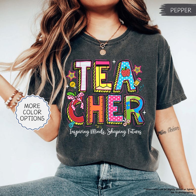 May include: A dark grey t-shirt featuring the word "TEACHER" in vibrant, block letters. The design incorporates an apple, stars, and school supplies. The text "Inspiring Minds, Shaping Futures" is displayed beneath the word.
