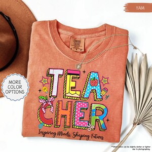 May include: A yam-coloured t-shirt with the word "TEACHER" in colourful, block letters. The design includes an apple, pencil, and stars. The text "Inspiring Minds, Shaping Futures" is below the word "TEACHER". A gold necklace is draped on the shirt.