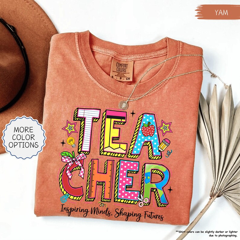 May include: A yam-coloured t-shirt with the word "TEACHER" in colourful, block letters. The design includes an apple, pencil, and stars. The text "Inspiring Minds, Shaping Futures" is below the word "TEACHER". A gold necklace is draped on the shirt.