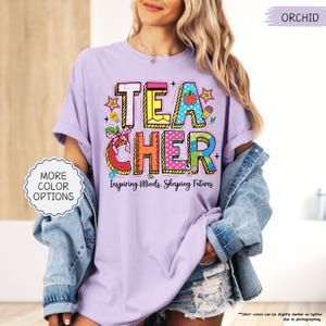 May include: Lilac-coloured t-shirt featuring the word "TEACHER" in vibrant, block letters. The design incorporates a strawberry, pencil, and ruler. The text "Inspiring Minds, Shaping Futures" is below the word "TEACHER".