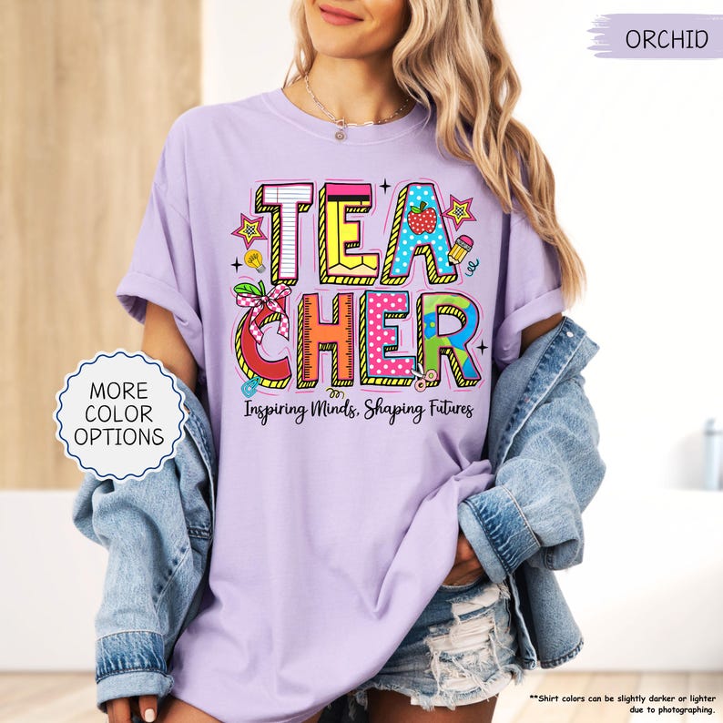 May include: Lilac-coloured t-shirt featuring the word "TEACHER" in vibrant, block letters. The design incorporates a strawberry, pencil, and ruler. The text "Inspiring Minds, Shaping Futures" is below the word "TEACHER".