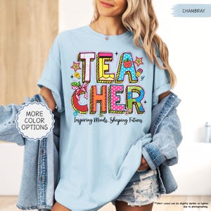 May include: Pale blue t-shirt with the word "TEACHER" in colourful, block letters. The shirt features a graphic design with school-themed elements and the text "Inspiring Minds, Shaping Futures". The shirt is part of a teacher-themed collection.