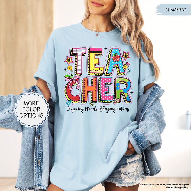 May include: Pale blue t-shirt with the word "TEACHER" in colourful, block letters. The shirt features a graphic design with school-themed elements and the text "Inspiring Minds, Shaping Futures". The shirt is part of a teacher-themed collection.