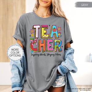 May include: A grey t-shirt featuring the word "TEACHER" in a colourful design with an apple, pencil, and stars. The text "Inspiring Minds, Shaping Futures" is below. The shirt is worn with a denim jacket and has rolled-up sleeves.