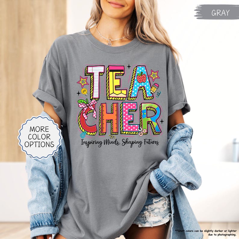 May include: A grey t-shirt featuring the word "TEACHER" in a colourful design with an apple, pencil, and stars. The text "Inspiring Minds, Shaping Futures" is below. The shirt is worn with a denim jacket and has rolled-up sleeves.