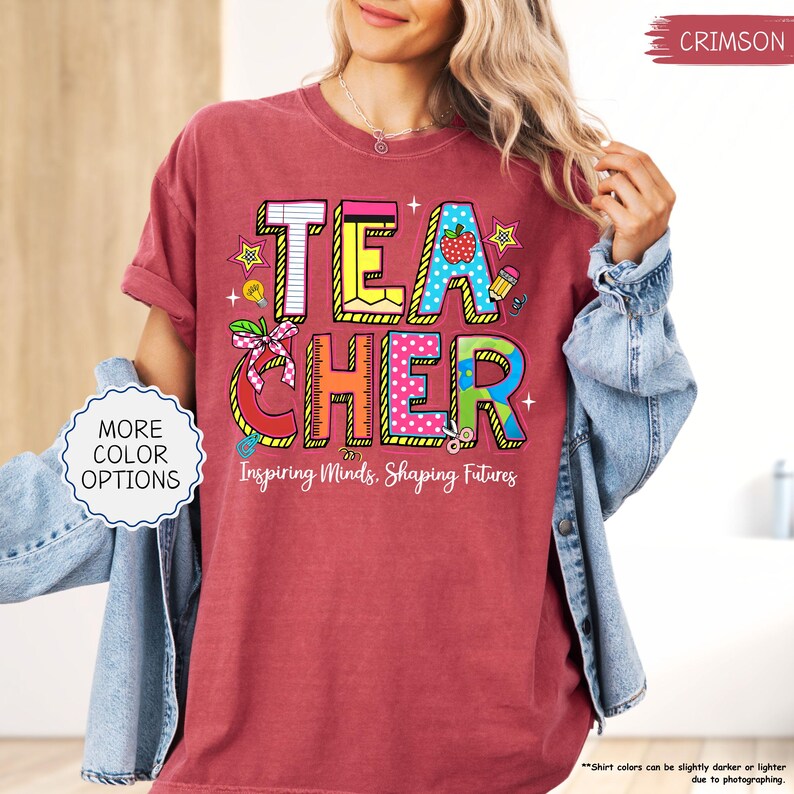 May include: Crimson t-shirt featuring the word "TEACHER" in colourful, block letters. The design includes an apple, pencil, and stars. The text "Inspiring Minds, Shaping Futures" is below the word "TEACHER".