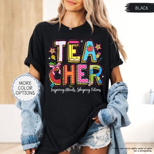 May include: Black t-shirt featuring the word "TEACHER" in vibrant, block letters. The design incorporates a strawberry, pencil, scissors, and the text "Inspiring Minds, Shaping Futures". This is a piece of clothing.