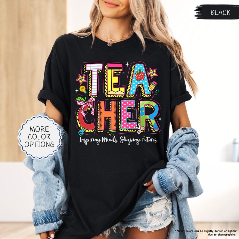 May include: Black t-shirt featuring the word "TEACHER" in vibrant, block letters. The design incorporates a strawberry, pencil, scissors, and the text "Inspiring Minds, Shaping Futures". This is a piece of clothing.