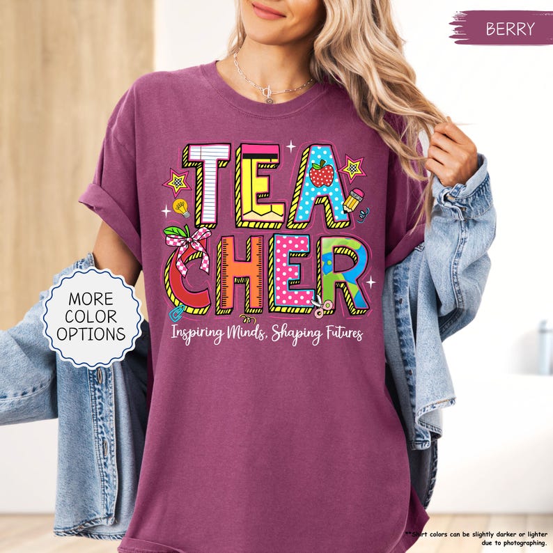 May include: A berry-coloured t-shirt featuring the word "TEACHER" in a vibrant, playful design. The design incorporates a strawberry, stars, and a pencil. The text "Inspiring Minds, Shaping Futures" is below the word "TEACHER".