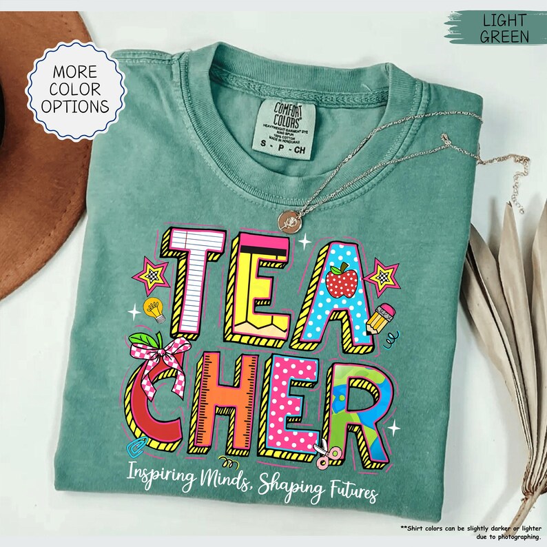 May include: A light green Comfort Colors t-shirt featuring the word "TEACHER" in a colourful, cartoon style. The design includes an apple, stars, and the text "Inspiring Minds, Shaping Futures". A silver necklace is draped across the shirt.