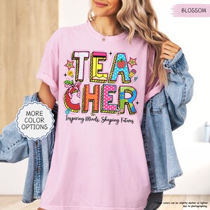 May include: A pale pink t-shirt featuring the word "TEACHER" in vibrant, block letters. The design incorporates a strawberry, pencil, and ruler. The text "Inspiring Minds, Shaping Futures" is below the word "TEACHER".