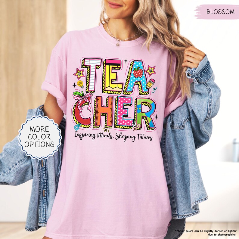 May include: A pale pink t-shirt featuring the word "TEACHER" in vibrant, block letters. The design incorporates a strawberry, pencil, and ruler. The text "Inspiring Minds, Shaping Futures" is below the word "TEACHER".